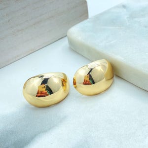 Può includere: Due orecchini dorati e arrotondati con una superficie lucida e riflettente. Gli orecchini hanno un design liscio e sono posti su uno sfondo bianco e azzurro. Sono gioielli.