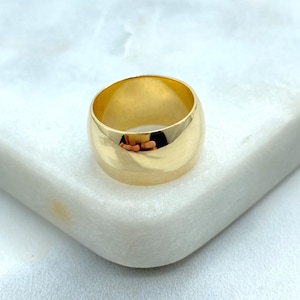 May include: A gold-colored ring with a wide band. The ring has a smooth, polished surface and reflects the surrounding light. The ring is sitting on a white surface.