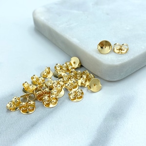 May include: A pile of gold-toned earring backs, some with a post and others with a butterfly closure. The earring backs are on a white surface.