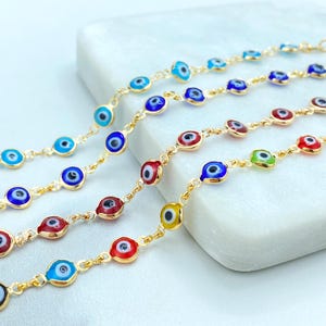 May include: Five gold chains with evil eye charms in different colors, including blue, red, green, and yellow. The chains are laid out on a white surface.