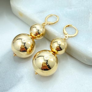 Puede incluir: Un par de pendientes colgantes dorados. Cada pendiente presenta dos esferas pulidas de diferentes tamaños, conectadas por un pequeño aro dorado. Los pendientes reflejan la luz, creando una apariencia brillante. Las joyas están sobre un fondo blanco y gris claro.