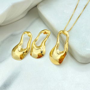 以下が含まれることがあります： ゴールドカラーのジュエリーセット。ペンダントネックレスと、お揃いのイヤリングが含まれています。ペンダントとイヤリングは、中が空洞になったユニークな有機的な形をしています。ジュエリーは白い表面に展示されており、光を反射して金の輝きを際立たせています。