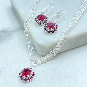 May include: A silver necklace and earring set featuring large, round, red gemstones surrounded by smaller, dark red stones. The necklace has a pendant with a matching gemstone, and the earrings have lever-back closures. The jewelry is displayed on a white surface.