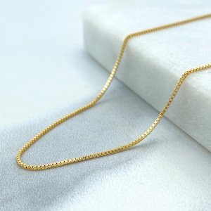 Può includere: Una collana a catena d'oro con un design a catena a scatola. La catena è appoggiata su una superficie bianca.