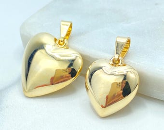 May include: Two gold-colored heart pendants with a polished, reflective surface. Each pendant has a loop at the top for attaching to a chain. The hearts are smooth and rounded, reflecting the surrounding environment. The pendants are likely jewelry.