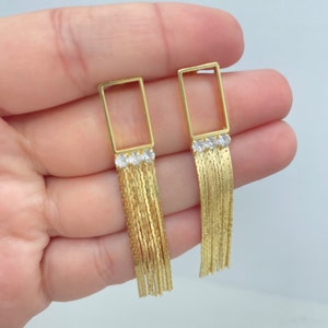 May include: Gold dangle earrings with a rectangular frame and a chain fringe. The frame is adorned with three small clear cubic zirconia stones.