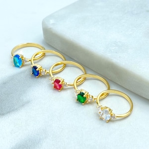 May include: Five gold-colored rings with oval-shaped gemstones in blue, dark blue, red, green, and clear. Each ring has a small cluster of three accent stones on either side of the main gemstone. The rings are displayed on a white surface.