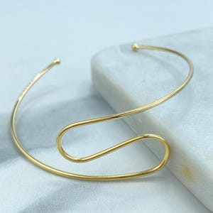 May include: A gold-colored, open cuff bracelet with a unique, flowing design. The bracelet features a slender, curved band that wraps around the wrist with a looped central element. The ends of the bracelet have small, rounded tips.