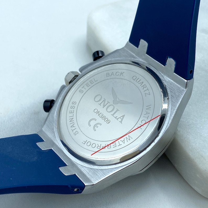 May include: The back of a silver watch with a blue silicone band. The watch face says "ONOLA" and "WATERPROOF" in red lettering. The watch is a quartz watch with a stainless steel back.