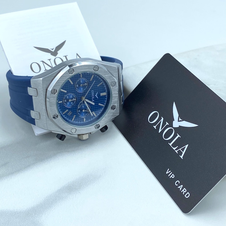 May include: A silver chronograph wristwatch with a blue face and a blue silicone strap. The watch has a silver case and a silver bezel with screws. The watch is on a white surface with a black card that says "ONOLA VIP CARD" in white text.
