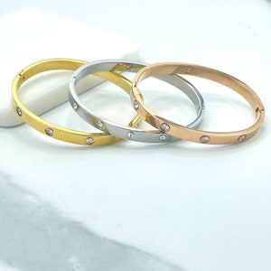 May include: Three metal bangles in gold, silver, and rose gold. Each bangle features small, clear gemstones evenly spaced around the band. The bracelets are displayed on a white surface, showcasing their polished finish and design.