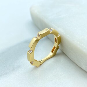 Könnte beinhalten: Ein goldfarbener Ring mit geometrischem Design. Der Ring hat rechteckige und quadratische Formen, mit kleinen, klaren Edelsteinen im Design. Der Ring wird auf einer weißen Marmoroberfläche präsentiert.