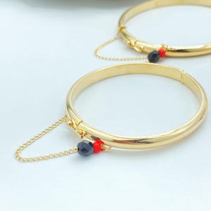 Puede incluir: Dos brazaletes de metal en tono dorado, cada uno con una fina cadena dorada unida a la banda. Una cuenta negra facetada y una pequeña cuenta roja redonda están situadas donde la cadena se conecta al marco circular y liso del brazalete.
