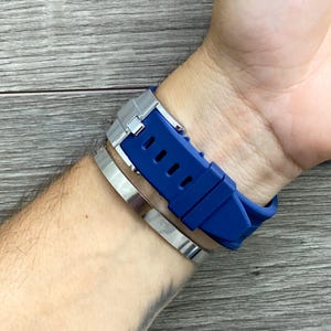 May include: A blue silicone watch band with a silver buckle. The band is on a person's wrist.
