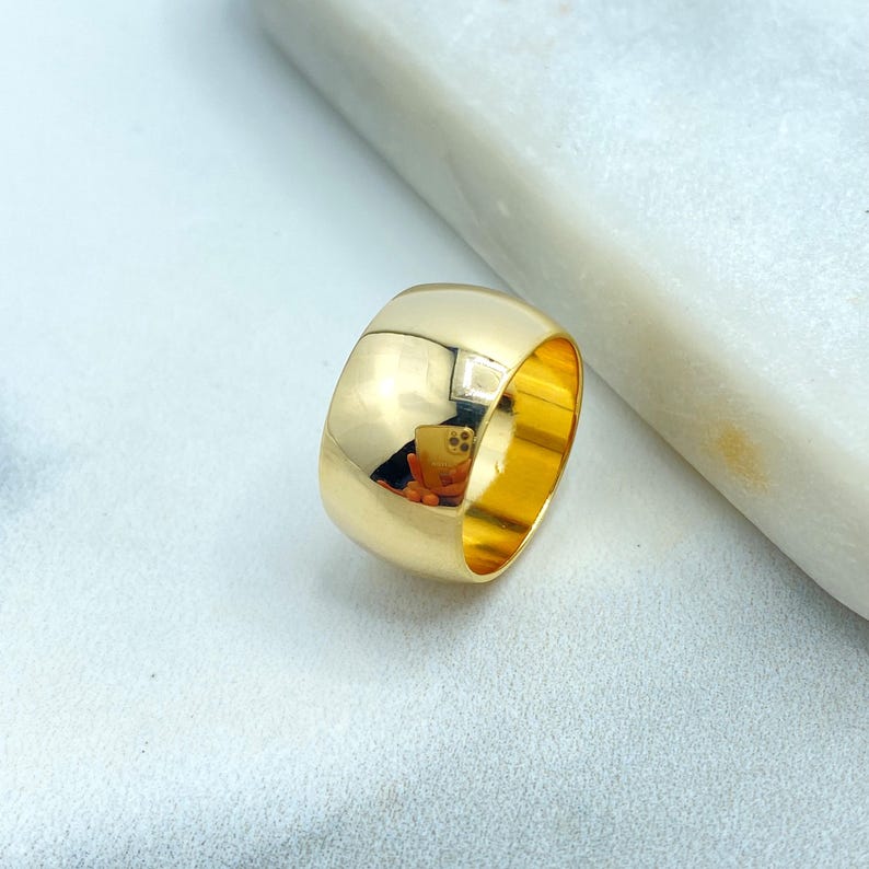 May include: A wide, gold-colored ring with a smooth, polished surface. The ring reflects the surrounding environment, showing a distorted image. The ring is set against a white and gray background.