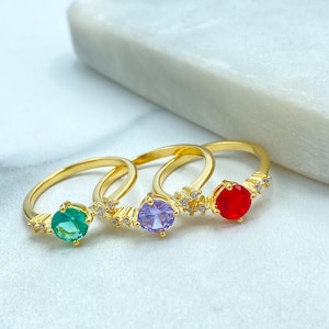 May include: Three gold rings with colorful gemstones. One ring features a green gemstone, another a purple gemstone, and the third a red gemstone. Each ring has small, clear accent stones.