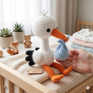 Puede incluir: Una cigüeña blanca de ganchillo hecha a mano con pico y patas naranjas, sosteniendo un bulto azul. La cigüeña tiene alas negras y está sentada sobre una manta beige. La etiqueta dice "Crochet by Adeeb".