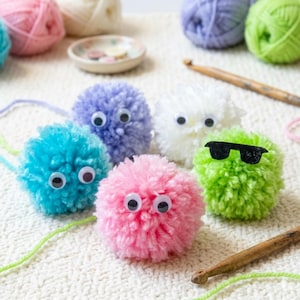 Peut inclure: Plusieurs pompons de fil colorés avec des yeux globuleux, dont bleu, violet, rose, blanc et vert. Un pompon vert porte des lunettes de soleil noires. Des crochets et des pelotes de laine sont en arrière-plan.