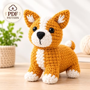 Op de afbeelding: Een gehaakte amigurumi-hond van oranje en wit garen, met zwarte plastic ogen, een zwart geborduurde neus en puntige oren. Een cirkelvormige badge in de linkerbovenhoek toont de tekst PDF PATTERN met een downloadpictogram erboven.