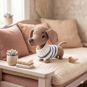 Puede incluir: Un perro salchicha de ganchillo con un cuello a rayas blancas y azules. El perro es marrón con una nariz negra y está sobre una superficie de punto color crema. Un pequeño pájaro de madera y libros en miniatura están cerca.