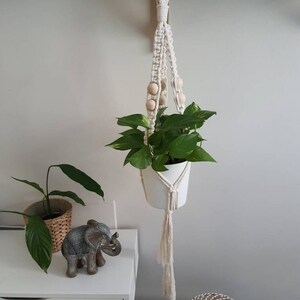 Può includere: Un portavasi in macramé bianco con perline di legno appeso a uno scaffale. Una pianta verde in vaso è nel portavasi. Il portavasi ha lunghi frange.