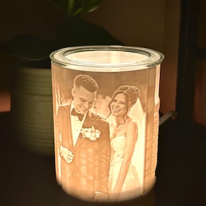 Peut inclure: Une lampe en céramique blanche avec une image 3D d'un couple dans un cadre de mariage. La lampe a une lueur chaude et est allumée.