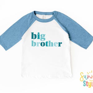 May include: White and blue raglan style t-shirt with the words "big brother" in teal and blue lettering. The shirt has three-quarter length sleeves and a crew neck. The Sunny Style logo is in the bottom right corner.