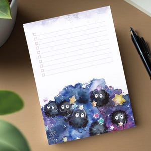 Könnte beinhalten: Ein Notizblock mit einer Aquarellillustration von Rußgeistern vor einem blau-violetten Galaxiehintergrund. Der Notizblock hat linierte Seiten mit Kontrollkästchen und ein schwarzer Stift liegt auf der Oberfläche.