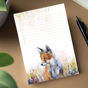 Könnte beinhalten: Notizblock mit einem Aquarellgemälde eines Fuchses. Der Fuchs hat orangefarbenes und weißes Fell und ist vor dem Hintergrund von hohem Gras platziert. Der Notizblock hat einen linierten Abschnitt für Notizen und einen Kontrolllistenabschnitt.
