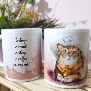 Könnte beinhalten: Zwei weiße Keramikbecher. Ein Becher zeigt den Text "Today: read, sleep, coffee, repeat" in schwarzer Schrift mit einem Aquarell-Hintergrund in Rosa und Braun. Der andere Becher hat eine Aquarell-Illustration einer Katze mit einer Zeitung.