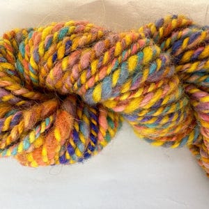 May include: A skein of colourful yarn with shades of yellow, orange, pink, blue, and purple. The yarn is twisted and fluffy, creating a textured look.