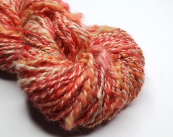 May include: A skein of yarn featuring a variegated blend of red, orange, pink, and cream fibres. The yarn is constructed from multiple twisted strands with a subtle metallic gold thread woven throughout, creating a textured and multi-tonal appearance.