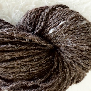 May include: A close-up of a ball of brown yarn with a textured appearance. The yarn is tightly wound, revealing the individual strands. The background is a soft, white, fluffy material. This yarn is suitable for knitting and crocheting projects.