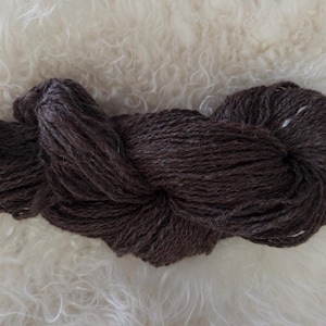May include: A skein of dark brown yarn, tightly wound, rests on a fluffy, white surface. The yarn appears to be a natural fiber, with a slightly textured appearance. The color is a rich, earthy tone, suitable for knitting or crocheting projects.
