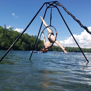 May include: A person performing aerial acrobatics on a black aerial hoop suspended from a tripod stand in a lake. The hoop is suspended over the water and the person is upside down in the hoop.