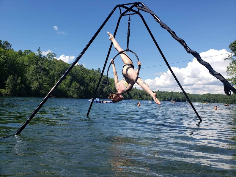 May include: A person performing aerial acrobatics on a black aerial hoop suspended from a tripod stand in a lake. The hoop is suspended over the water and the person is upside down in the hoop.