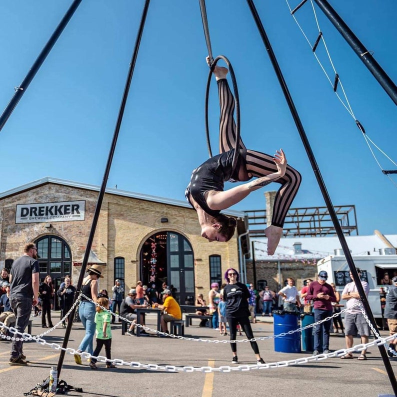 May include: A woman performs an aerial silks routine on a black and white striped fabric hanging from a metal frame. The silks are suspended from a metal frame with a black and white striped fabric. The woman is wearing a black leotard with a black and white striped pattern. The background is a brick building with a sign that reads "Drekker Brewing Company".