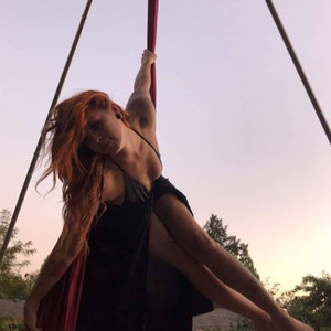 May include: A woman in a black dress is performing aerial silks. She is hanging upside down from a red silk fabric suspended from a metal frame. The background is a sunset sky.