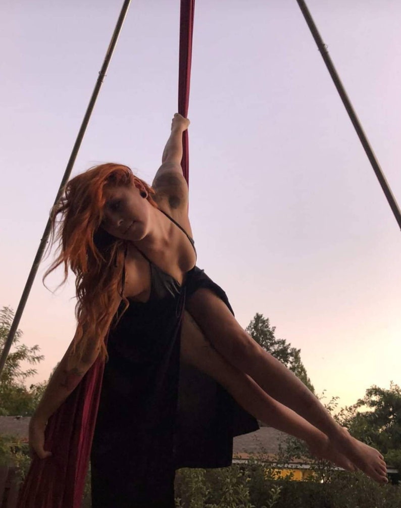 May include: A woman in a black dress is performing aerial silks. She is hanging upside down from a red silk fabric suspended from a metal frame. The background is a sunset sky.