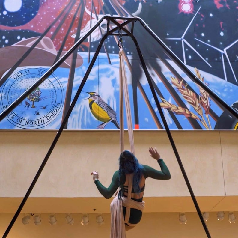 May include: A person in a green and black outfit is suspended in the air by a white fabric hammock. The hammock is attached to a black metal frame. The person has blue hair and is holding onto the fabric. A yellow bird is painted on a wall behind the person.