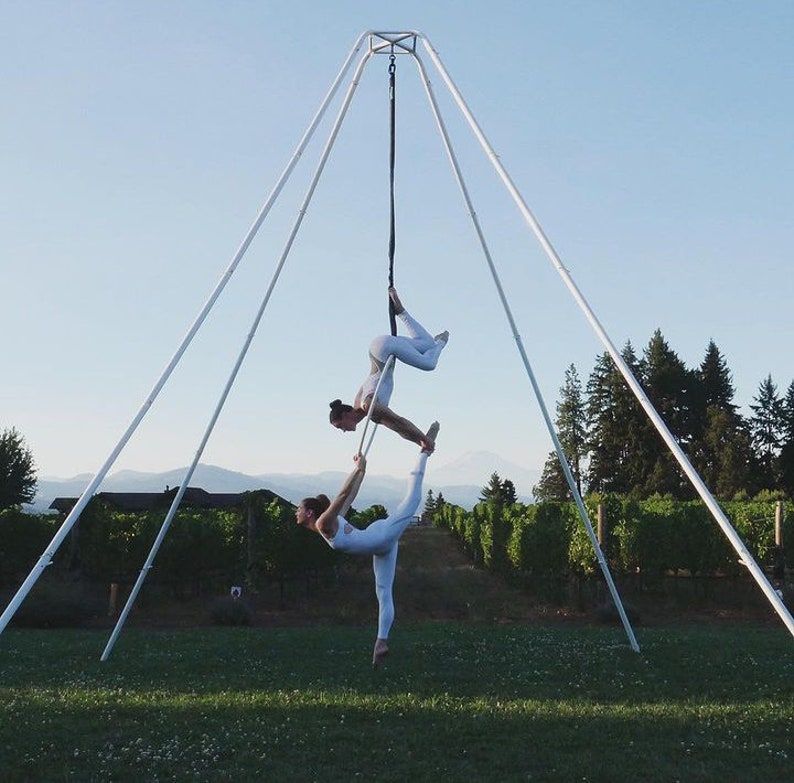 May include: Two people in white clothing perform an aerial silks routine on a white aerial rig outdoors. The rig is set up in a vineyard with green grass and rows of grapevines.