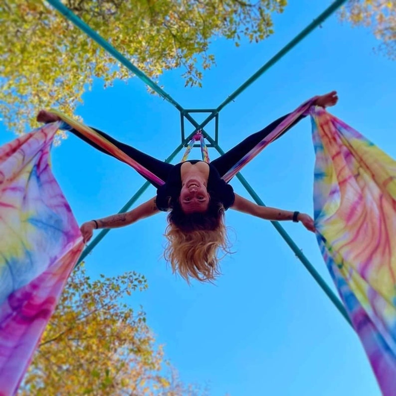 May include: A person performing aerial silks, hanging upside down from a colorful fabric suspended from a metal frame. The silks are a vibrant rainbow tie-dye pattern, and the person is smiling.