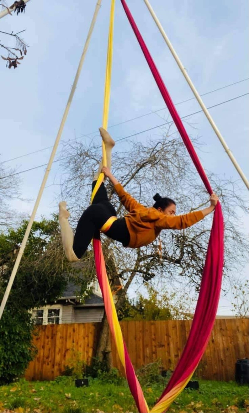 May include: A woman performs aerial silks in a backyard. She is wearing a brown sweater and black leggings. The silks are yellow, pink, and red.