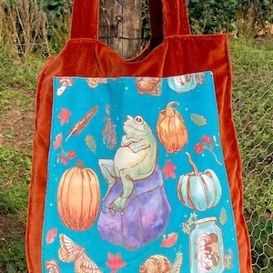 May include: A bright orange velvet tote bag with a blue fabric lining featuring a whimsical illustration of a frog, pumpkins, mushrooms, a snail, and a jar of mushrooms.