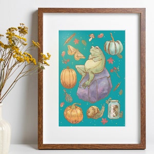 Könnte beinhalten: Eine gerahmte Illustration eines grünen Frosches, der auf einem violetten Felsen sitzt, vor einem blaugrünen Hintergrund. Der Frosch ist von verschiedenen herbstlichen Elementen umgeben, darunter Kürbisse, Blätter, Pilze und ein Glas mit Pilzen.