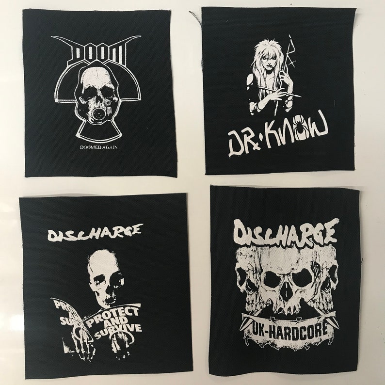 May include: Four black and white iron-on patches. The top left patch features a skull with a gas mask and the text "DOOM DOOMED AGAIN". The top right patch features a woman with long hair and the text "DR. KNOW". The bottom left patch features a man with a skull face and the text "DISCHARGE PROTECT AND SURVIVE". The bottom right patch features three skulls with the text "DISCHARGE UK-HARDCORE".