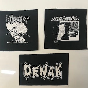 May include: Three black fabric patches with white text and graphics. The first patch has the text "DISRUPT" and a graphic of a person wearing a cowboy hat and a clown mask. The text "RID THE CANCER" is below the graphic. The second patch has the text "SLEEP" and a graphic of a person's hand and a circular design. The third patch has the text "DENAK" in a stylized font.