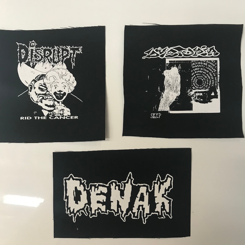 May include: Three black fabric patches with white text and graphics. The first patch has the text "DISRUPT" and a graphic of a person wearing a cowboy hat and a clown mask. The text "RID THE CANCER" is below the graphic. The second patch has the text "SLEEP" and a graphic of a person's hand and a circular design. The third patch has the text "DENAK" in a stylized font.