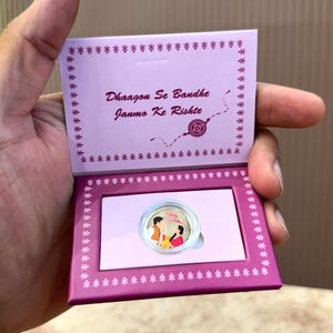 Puede incluir: Una caja de regalo morada y rosa con el texto "Dhaagon Se Bandhe Janmo Ke Rishte" y una pequeña moneda con una colorida ilustración de dos personas. La caja está abierta, revelando la moneda.
