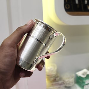May include: A silver-colored mug with a handle. The mug has a cylindrical body with a textured pattern near the top and a smooth, brushed finish on the rest of the body. The handle is curved and attached to the side of the mug.